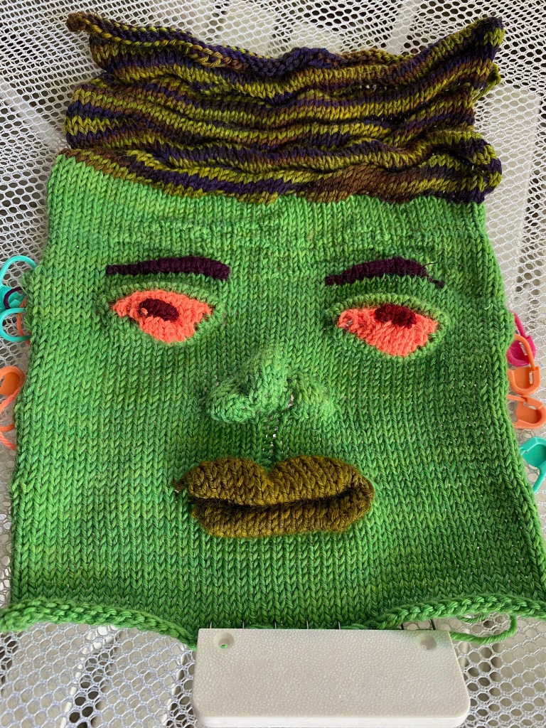 Outward facing side of green machine knit face with markers on the sides to indicate row count