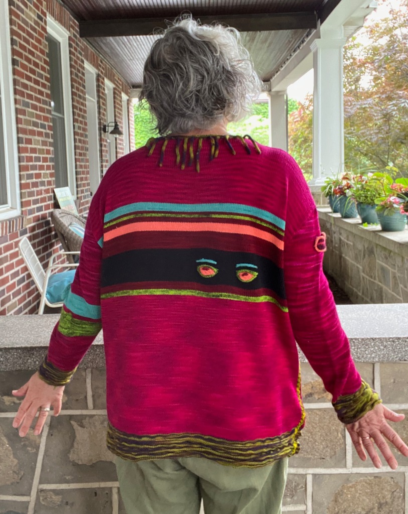 Back of machine knit sweater with eyes looking out of a black stripe and purple and green cords around the neck