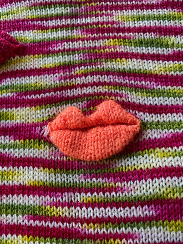 Light pink-orange machine-knit lips on a background of pink, green, and white knitted in narrow partial stripes, front side of work